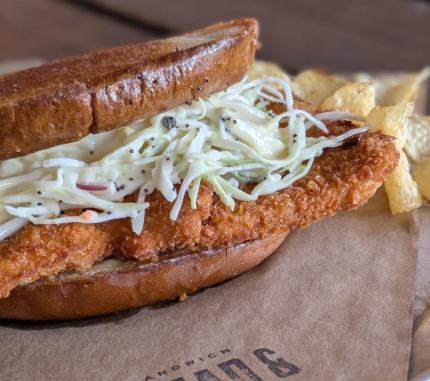 A breaded pork sandwich topped with slaw beside chips at Bread & Circus Kitchen