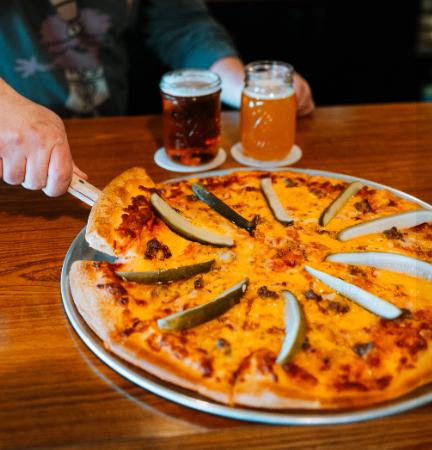 A pizza topped with dill pickles being sliced at a table with two glasses of beer at Charlie's Pizza