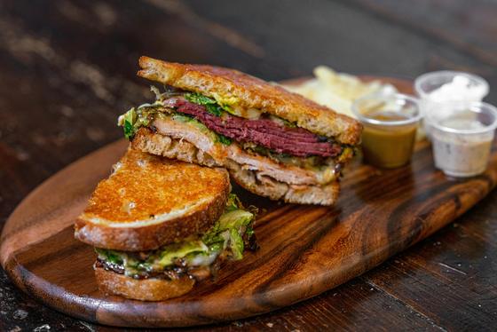 A toasted deli sandwich cut in half on a wooden board with dipping sauces